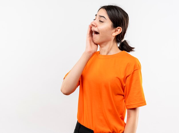 Niña vistiendo camiseta naranja con aspecto cansado quiere dormir bostezando de pie sobre la pared blanca