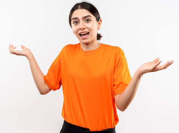 Niña vistiendo camiseta naranja confundido encogiéndose de hombros sin respuesta de pie sobre la pared blanca