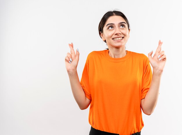 Niña vistiendo camiseta naranja haciendo deseo deseable cruzar los dedos feliz y alegre de pie sobre la pared blanca