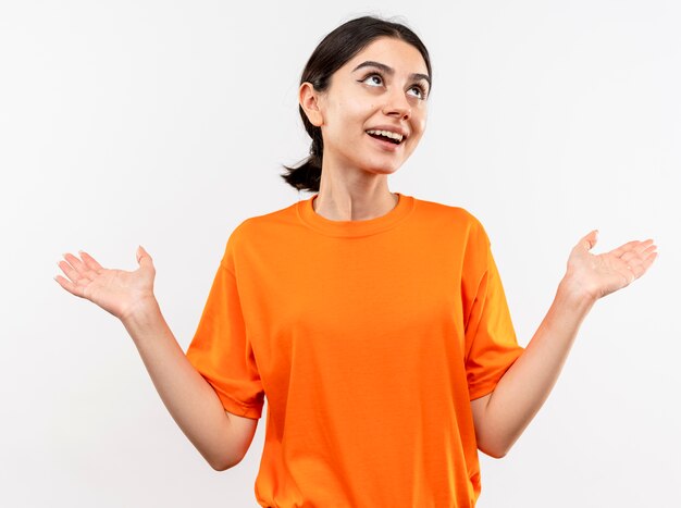 Niña vistiendo camiseta naranja mirando hacia arriba siendo confundido y feliz extendiendo los brazos a los lados sonriendo de pie sobre la pared blanca