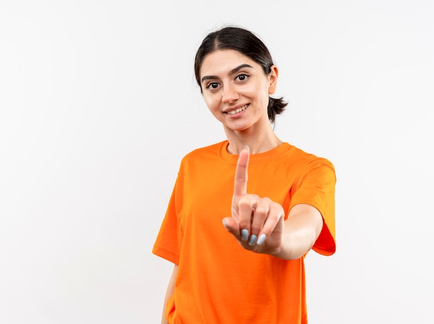 Niña vistiendo camiseta naranja mostrando el dedo índice con una sonrisa en la cara de pie sobre la pared blanca