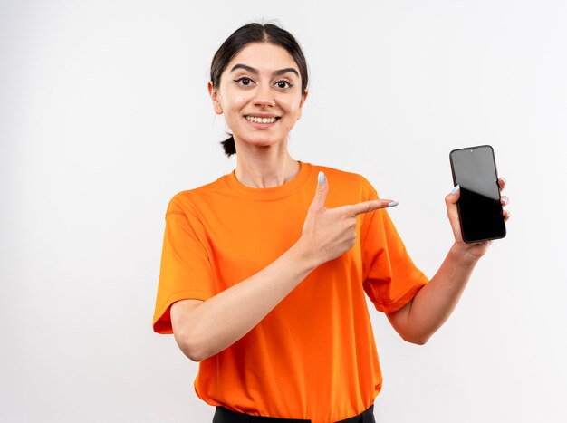 Niña vistiendo camiseta naranja mostrando smartphone apuntando con el dedo índice sonriendo alegremente de pie sobre la pared blanca