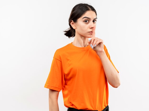 Niña vistiendo camiseta naranja con rostro serio haciendo gesto de silencio con el dedo en los labios de pie sobre la pared blanca