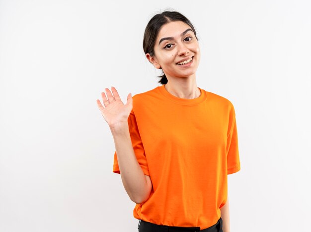 Niña vistiendo camiseta naranja sonriendo amistosamente saludando con la mano de pie sobre la pared blanca