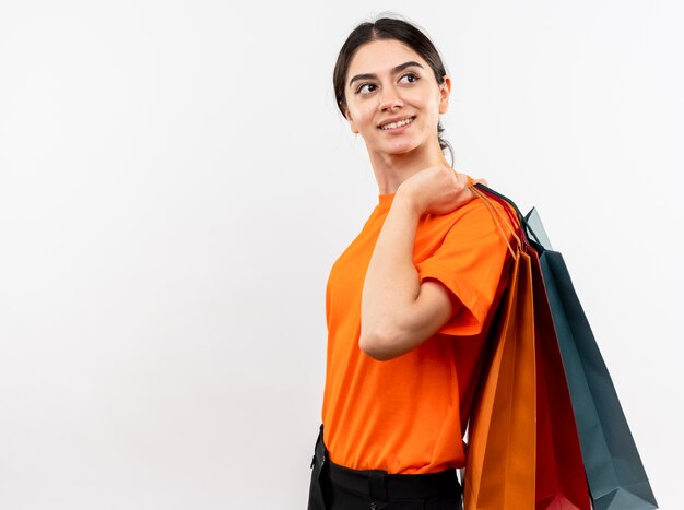 Niña vistiendo camiseta naranja sosteniendo bolsas de papel mirando a un lado con cara hapy sonriendo alegremente de pie sobre la pared blanca