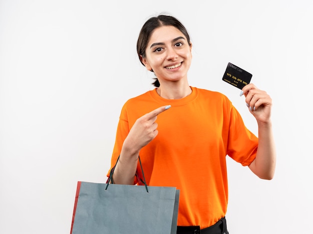 Foto gratuita niña vistiendo camiseta naranja sosteniendo bolsas de papel mostrando tarjeta de crédito apuntando con el dedo sonriendo de pie sobre la pared blanca