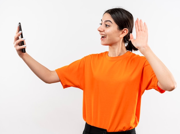 Niña vistiendo camiseta naranja con videollamada feliz y positivo sonriendo saludando con la mano de pie sobre la pared blanca