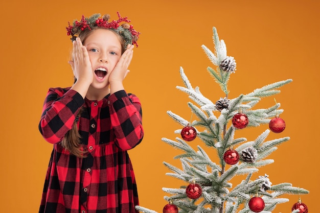 Niña vistiendo una corona de navidad en camisa a cuadros asombrado de pie junto a un árbol de navidad sobre la pared naranja