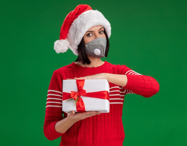 Niña vistiendo gorro de Papá Noel y máscara protectora con paquete de regalo aislado en la pared verde