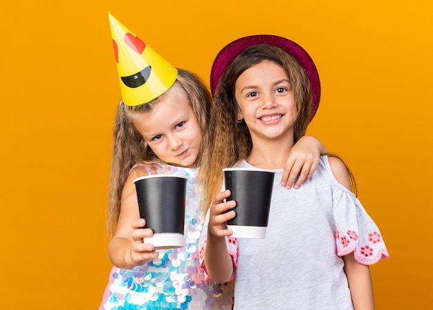 Niñas bonitas complacidas con sombreros de fiesta sosteniendo vasos de papel aislado en la pared naranja con espacio de copia