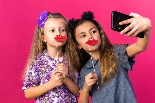 Niñas bonitas complacidas sosteniendo labios falsos en palos tomando selfie aislado en pared rosa con espacio de copia