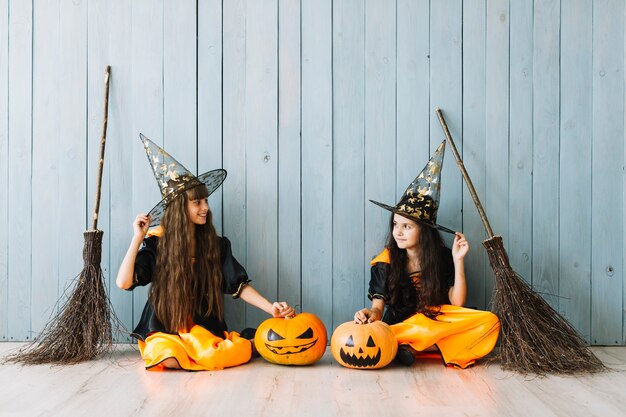 Niñas en disfraces de Halloween con sombreros puntiagudos y escobas sentados cerca