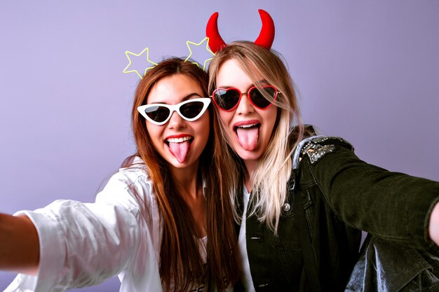 Niñas divertidas haciendo selfie, gafas vintage, diademas y cintas para el cabello de fiesta de estrellas, ropa juvenil informal, estado de ánimo positivo.