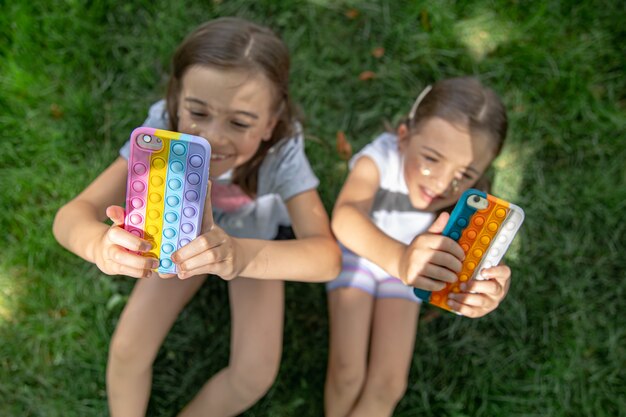 Niñas divertidas en la hierba con teléfonos en un estuche con espinillas, un juguete antiestrés de moda.