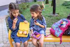 Foto gratuita niñas de la escuela sentadas en un banco en el patio de la escuela y comiendo de las loncheras.