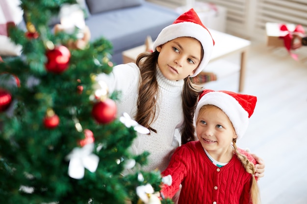 Niñas hermanas felices mirando el árbol de navidad decorado en el salón interior