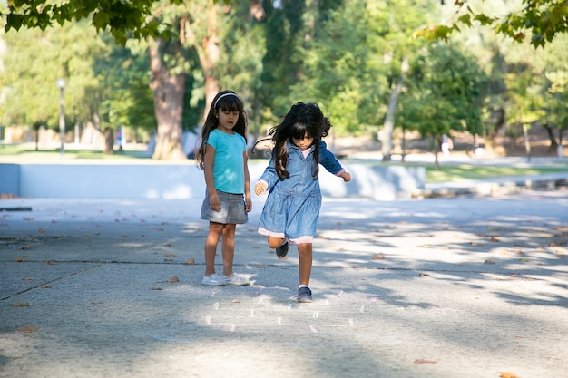 Niñas de pelo bastante negro jugando a la rayuela en el parque de la ciudad. Longitud total, espacio de copia. Concepto de infancia