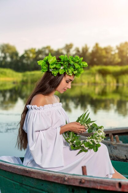 La ninfa de pelo largo y oscuro con un vestido vintage blanco sentada en un bote en medio del río. En el cabello una corona de hojas verdes con flores blancas. Sesión de fotos de fantasía.