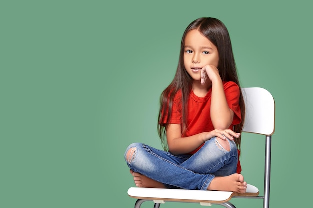 Foto gratuita niñita con camiseta roja y posando en una silla con fondo verde