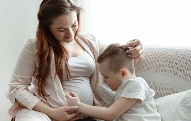 Niño abrazando el vientre de su madre embarazada en el sofá de casa.