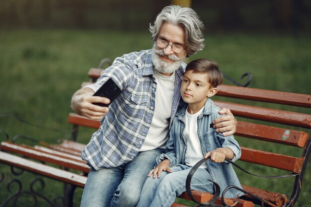 Niño y abuelo sentados en un banco. Familia en el parque. Anciano jugando con su nieto. El abuelo usa un teléfono.