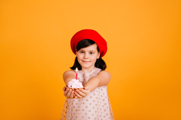 Niño adorable en elegante boina celebrando un cumpleaños. Niño de sexo femenino caucásico que sostiene la torta con la vela aislada en la pared amarilla.