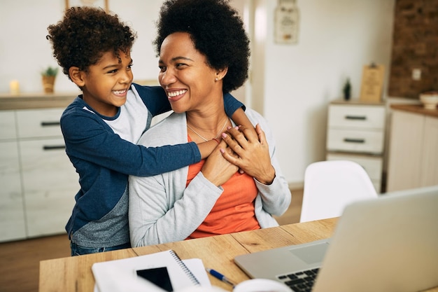 Niño afroamericano feliz abrazando a su madre que está trabajando en una laptop en casa