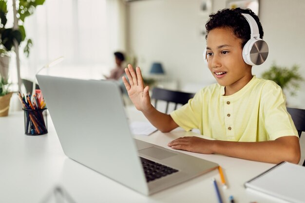 Niño afroamericano que usa una computadora portátil y saluda durante una videollamada mientras estudia en casa