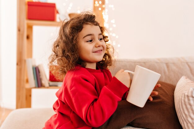 Niño alegre bebiendo té. Niño rizado encantador que sostiene la taza.