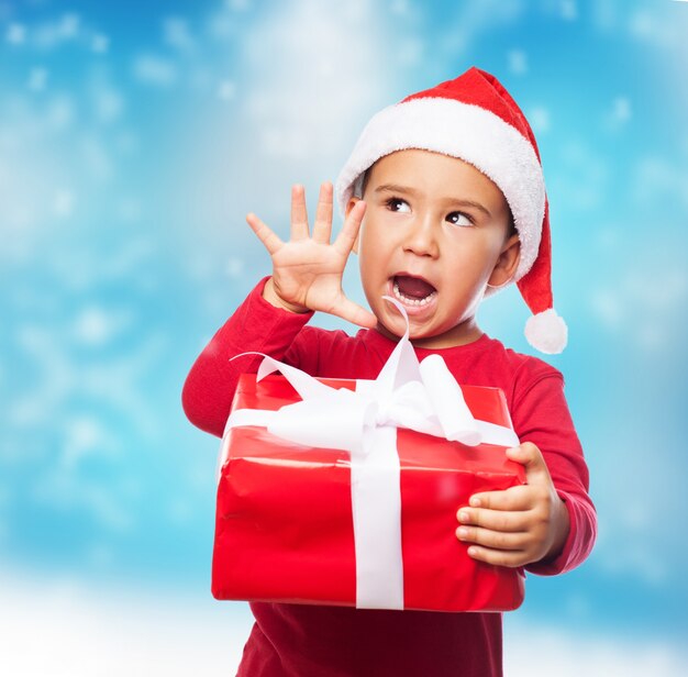 Niño alegre jugando con su mano derecha y sujetando un regalo