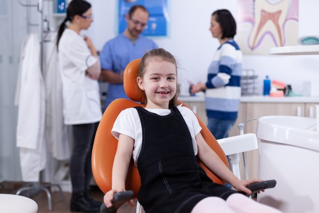 Niño alegre sentado en una silla en el consultorio del dentista durante la visita por mal tratamiento dental y los padres discutiendo con el médico. Niño con su madre durante el chequeo de dientes con estomatólogo sentado en una silla.