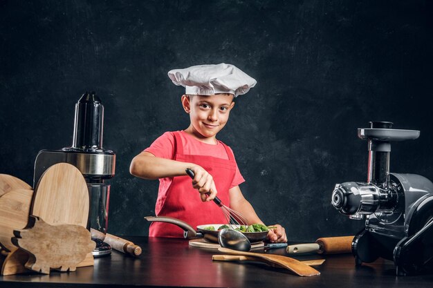 Un niño alegre con sombrero y delantal de chef está preparando verduras para cocinar.