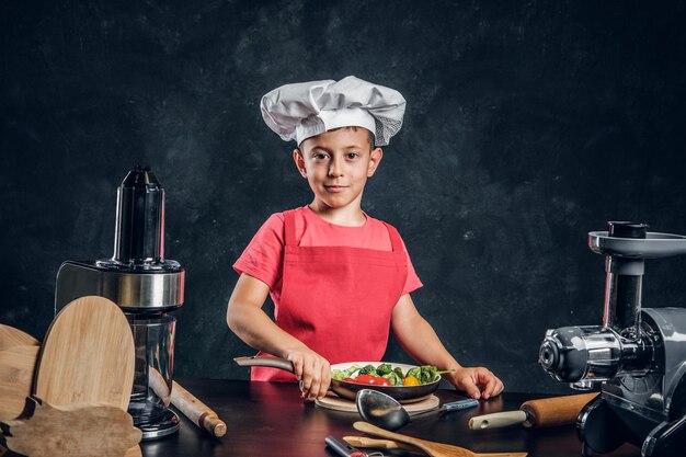 Un niño alegre con sombrero y delantal de chef está preparando verduras para cocinar.