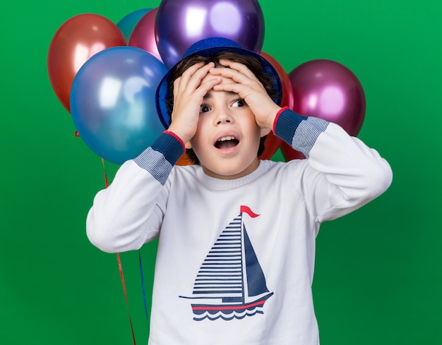 Foto gratuita niño arrepentido con sombrero de fiesta azul de pie delante de globos agarró la cabeza