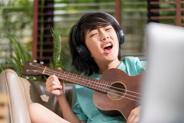 Niño asiático canta mientras aprende un curso de estudio de ukelele en línea con diversión y salida en casa. Estudio en línea concepto de quedarse en casa. Niño asiático tocando ukelele y cantando en la sala de estar en casa.