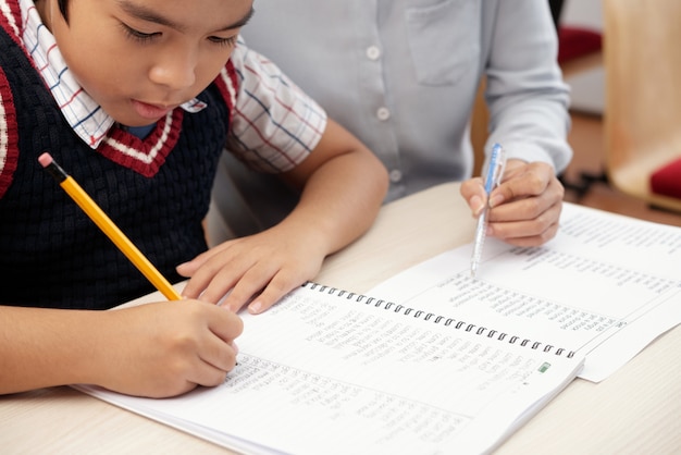 Niño asiático escribiendo en cuaderno y mujer irreconocible sentada y mirando