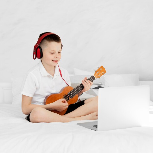 Niño con auriculares tocando el ukelele y sentado en la cama