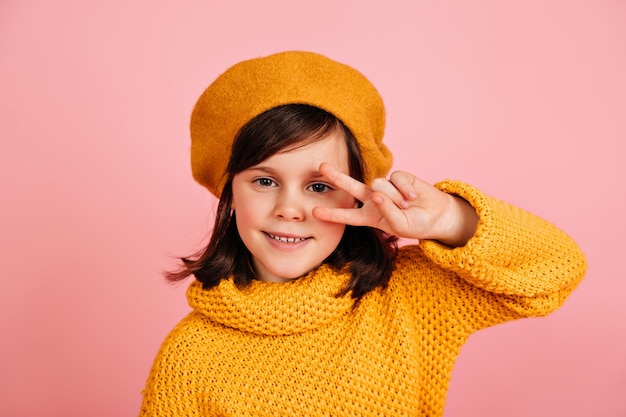 Niño en boina amarilla posando con el signo de la paz Foto de estudio de niña aislada sobre fondo rosa