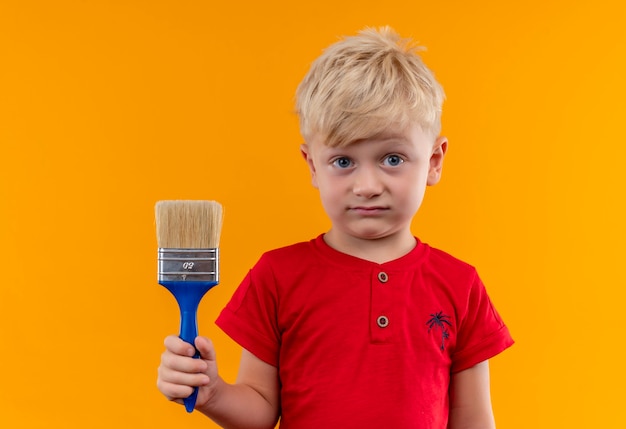 Foto gratuita un niño bonito con cabello rubio y ojos azules vistiendo camiseta roja sosteniendo un pincel azul mirando en una pared amarilla