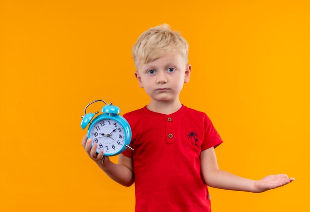 Un niño con cabello rubio y ojos azules con camiseta roja sosteniendo un reloj despertador azul mientras mira en una pared amarilla