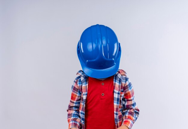 Un niño con cabello rubio vistiendo camisa a cuadros cerrando su rostro con casco azul sobre una pared blanca