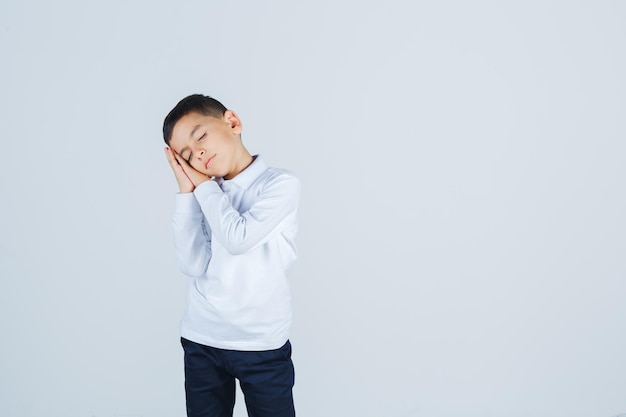 Niño con camisa blanca, pantalones apoyados en las palmas de las manos como almohada y con sueño, vista frontal.