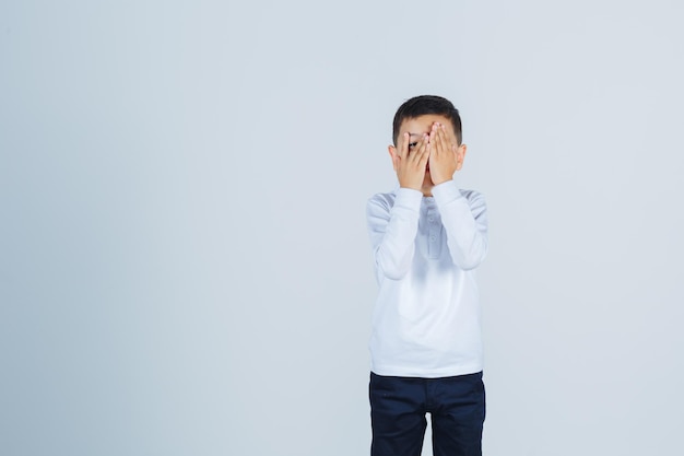 Niño con camisa blanca, pantalones mirando a través de los dedos y mirando asustado, vista frontal.