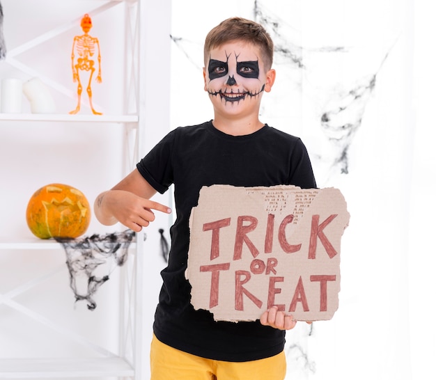 Niño con cara pintada con cartel de truco o trato para halloween