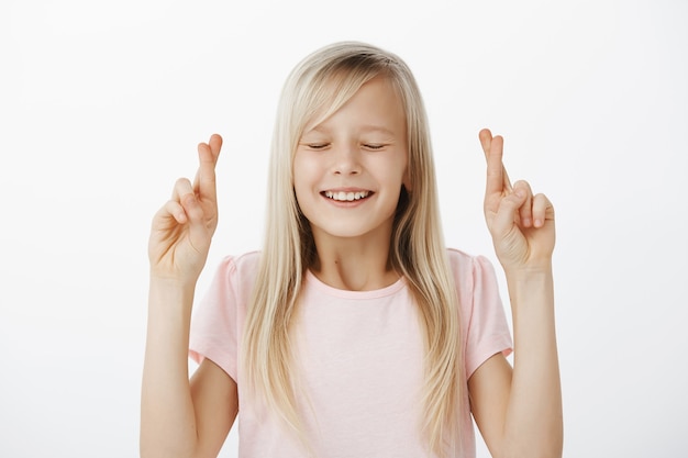 Foto gratuita niño caucásico fiel positivo con cabello rubio en camiseta rosa, cerrando los ojos y sonriendo con esperanza, cruzando los dedos índices mientras pide un deseo, quiere algo y le pide a dios que cumpla el sueño