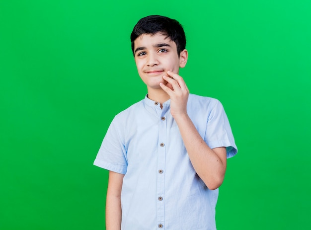 Niño caucásico joven complacido tocando la barbilla aislada en la pared verde con espacio de copia