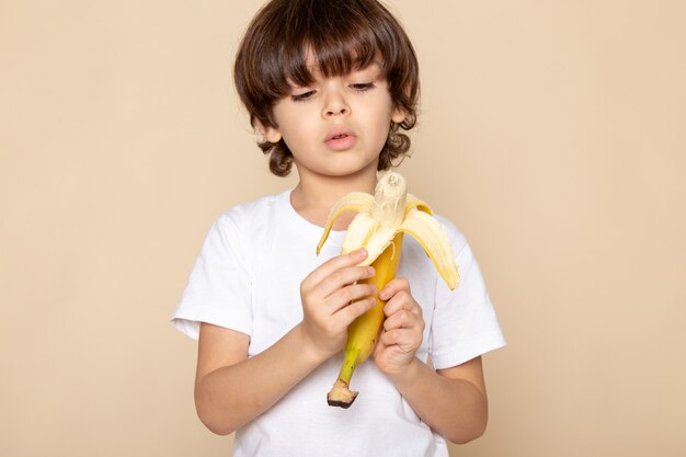 niño chico lindo dulce en camiseta blanca pelando plátano en pared rosa