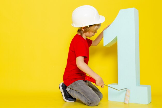 niño chico rubio pelo lindo adrorable en camiseta roja y figura de número de pintura de casco blanco sobre amarillo