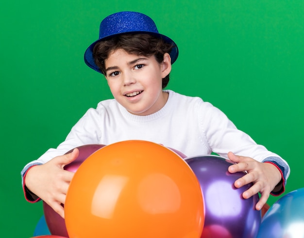 Niño contento con sombrero de fiesta azul de pie detrás de globos aislados en la pared verde