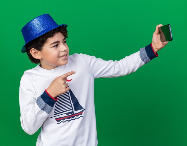 Niño contento con sombrero de fiesta azul tomar un selfie puntos en el frente aislado en la pared verde